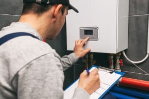 A technician in a cap and overalls adjusts the settings on a wall-mounted control panel, likely for a water heater installation in Plymouth County MA. He holds a clipboard with papers in one hand and uses the other to press buttons on the panel, showcasing his skills as a plumbing contractor.