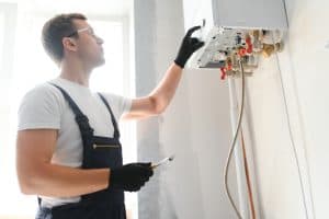A technician wearing gloves and goggles adjusts a boiler in a well-lit room in Plymouth County, MA. Holding a screwdriver, the plumber appears focused on the unit's controls mounted on the wall, conveying a sense of plumbing repairs and maintenance.