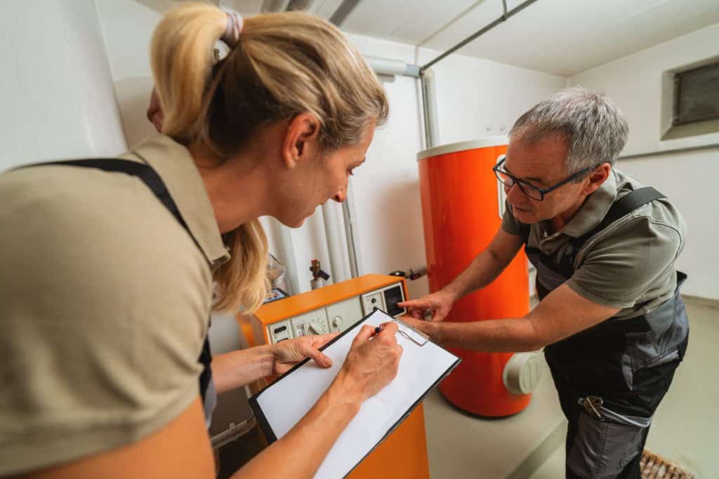 Two technicians in work uniforms check a heating system in a utility room; a plumber Plymouth expert takes notes on a clipboard while the other points to controls on an orange boiler tank.