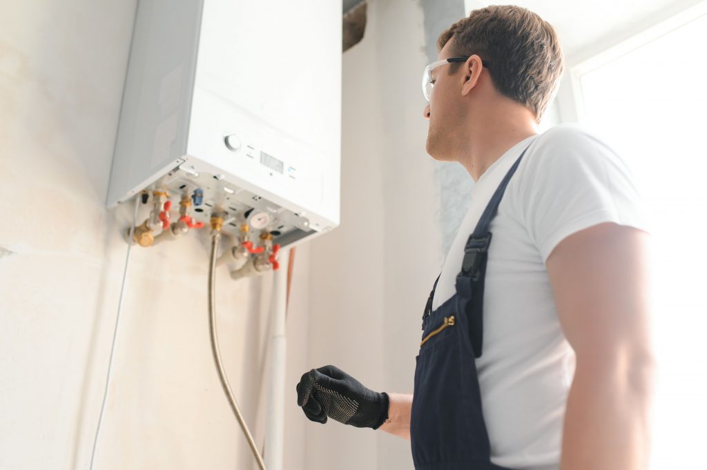 A technician wearing glasses, gloves, and overalls inspects a wall-mounted boiler or water heater in a bright, indoor setting—serving homes throughout Bristol and Middlesex counties.