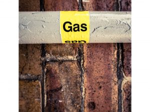 A gas pipe with a yellow label reading "Gas" is mounted on a wet brick wall in Plymouth County, MA. The horizontal pipe glistens with water droplets, reflecting the skill of local plumbers known for their expertise in plumbing repairs and installations.