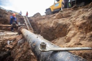 At a bustling construction site in Plymouth County, MA, a large metal pipe lies partially buried, hinting at ongoing plumbing repairs. A wrench rests on the pipe as workers in safety gear and a truck can be seen by the trench ladder, diligently managed by an expert plumbing contractor.