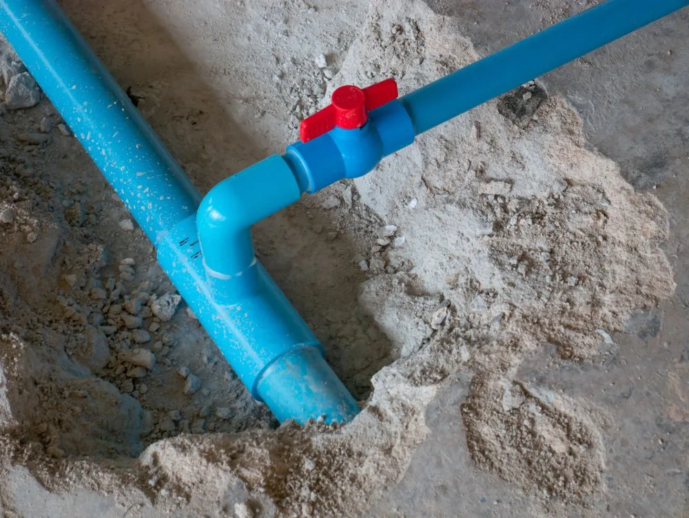 A blue PVC water pipe with a red valve handle runs through a concrete floor, exposed in a dug-out section surrounded by loose dirt and gravel—just the kind of repair handled by an experienced plumber in Plymouth, MA.