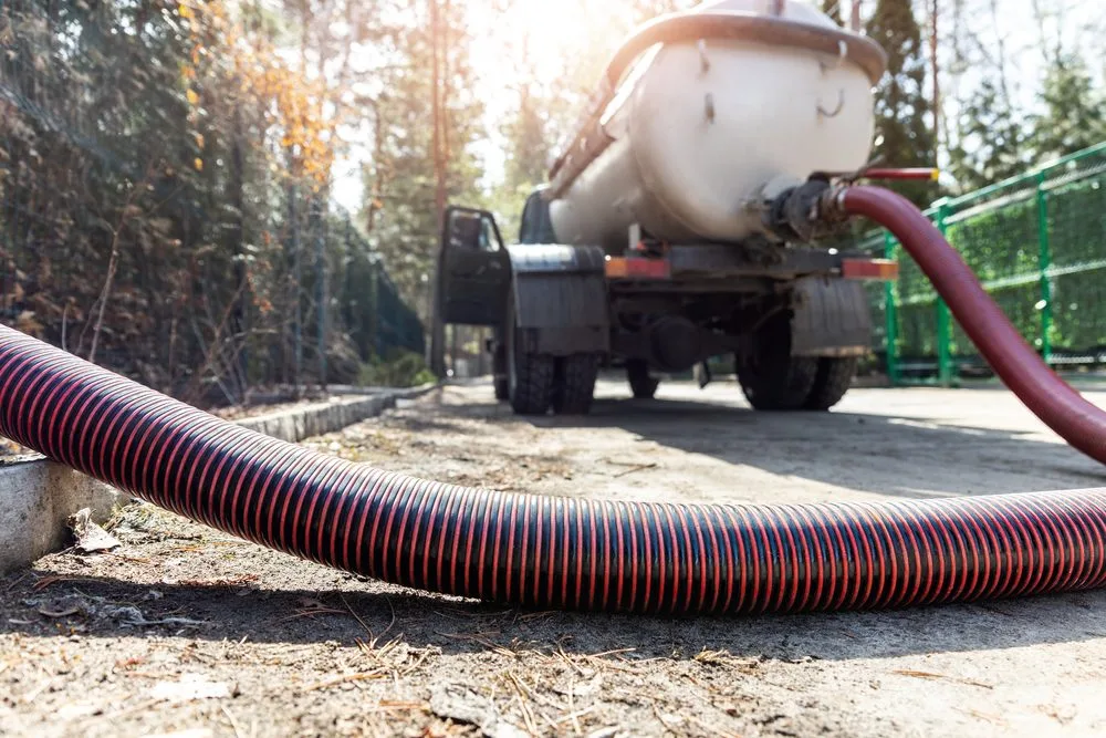 A large vacuum hose extends from a septic tank truck parked outdoors on a paved surface in Bristol and Middlesex County, with trees and a green fence in the background under bright sunlight.