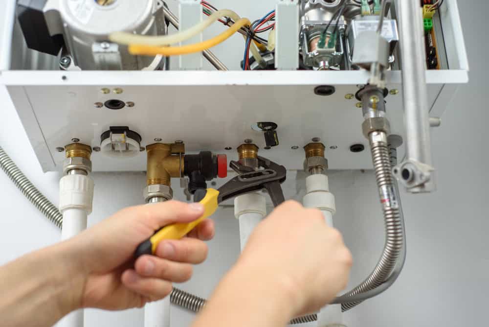 A plumber Plymouth, MA uses a wrench to tighten a pipe connection on a gas boiler, with various pipes and wires visible inside the boiler’s open panel.