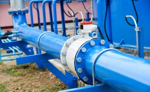 A large blue industrial pipeline with a white valve and a red cap is outdoors, surrounded by blue support structures and some foliage. This setup, likely part of a water or gas distribution system, showcases the expertise of a plumbing contractor in Plymouth County, MA.
