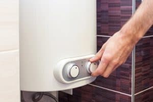 A person adjusts the temperature knob on a white water heater mounted on a wall with purple tile accents, showcasing the expertise of a Plumbing Contractor in Plymouth County, MA.