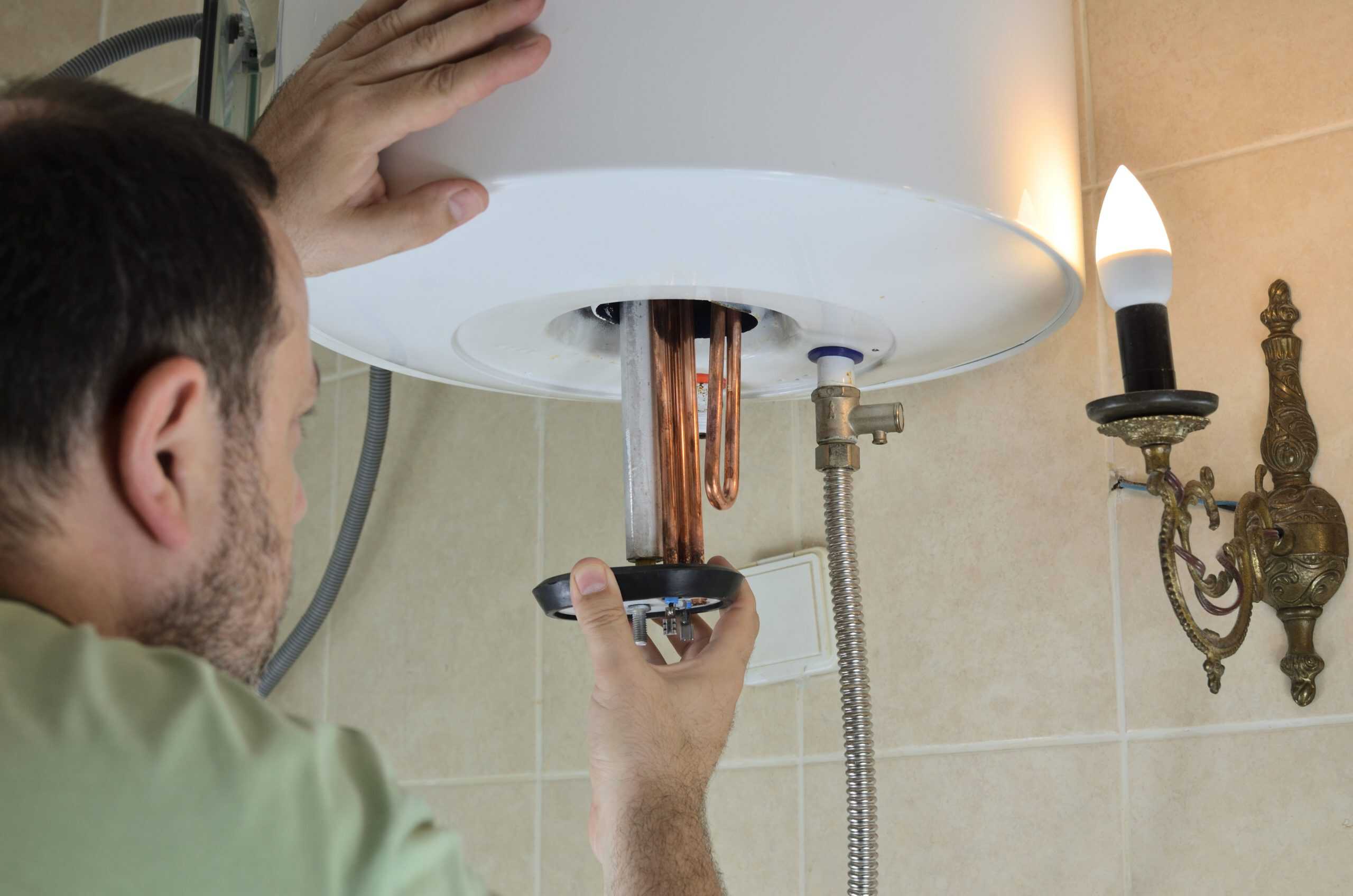 A plumber in Plymouth County, MA, is expertly inspecting or repairing a wall-mounted water heater in a bathroom. They are carefully adjusting a component inside the heater, under the glow of a nearby single-bulb light fixture on the tiled wall.