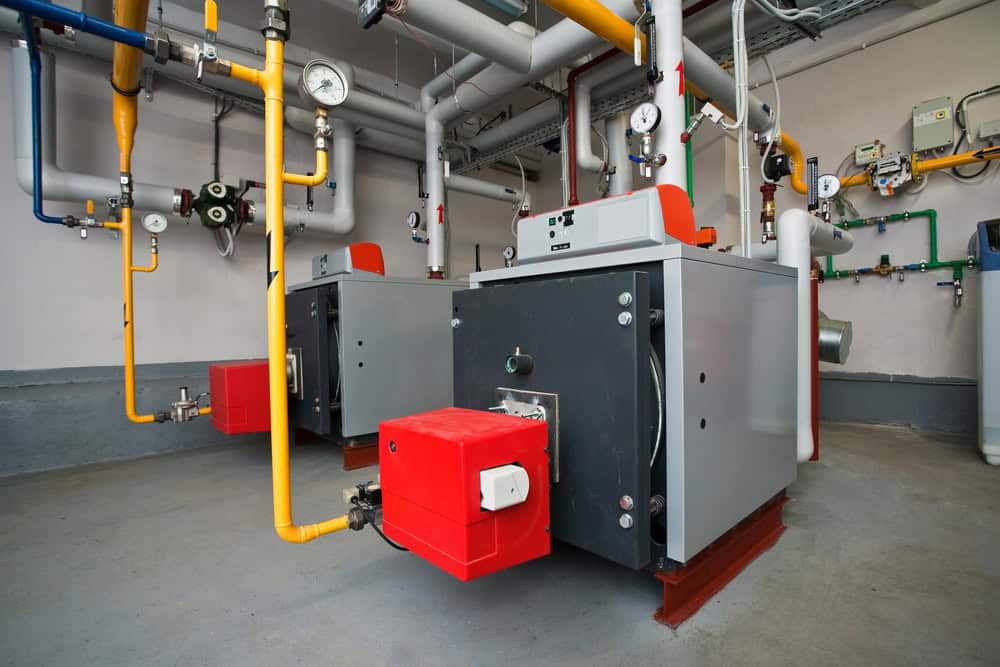 Two large industrial boilers with red burners are installed in a utility room in Middlesex, surrounded by yellow, green, and blue pipes, gauges, and control panels on the walls and ceiling.
