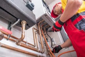 A plumber in red overalls and a cap repairs pipes under a boiler in Plymouth County, MA. He skillfully uses a tool to adjust the intricate copper plumbing, focusing on his precise actions indoors.