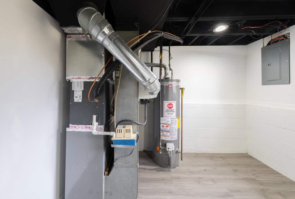 A basement utility room with exposed ductwork, a furnace, and a water heater showcases the expertise of a Plumbing Contractor in Plymouth County, MA. The light wood floor and white walls create a clean, bright atmosphere. A gray electrical panel is visible on the right wall.