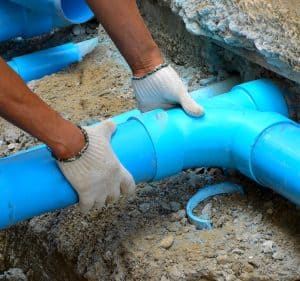 A person wearing white gloves is assembling blue plastic pipes in a sandy trench, showcasing the precision of a skilled plumbing contractor in Plymouth County, MA. The image focuses on the individual's hands expertly connecting the pipe sections.
