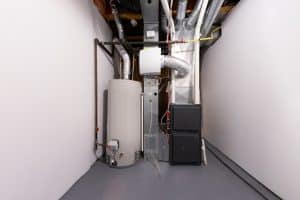 A basement in Plymouth County, MA, features a furnace system and expertly installed water heater. The white walls and gray floor complement the visible pipes and ducts overhead. This clean, well-organized space stands ready for any plumbing repairs by a skilled plumber in the area.