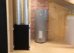 A modern HVAC unit with two large metal ducts beside a cylindrical metallic water heater, expertly installed by a plumbing contractor in Plymouth County MA, stands in a room featuring wooden walls and a brick accent wall under a sloped ceiling.