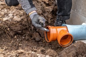 A person wearing gloves inspects a newly installed orange PVC pipe joint with a Y-fitting, partially buried in a trench amidst soil and rocks. The scene suggests ongoing plumbing work, possibly undertaken by a skilled plumbing contractor in Plymouth County, MA.