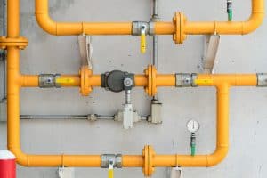 A network of yellow gas pipes, expertly installed by a top plumbing contractor in Plymouth County, MA, features various valves and connectors mounted on a gray wall. A pressure gauge is visible at the bottom right. The pipes run horizontally and vertically, creating an industrial grid pattern.