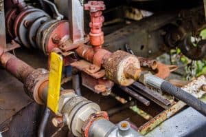 Close-up of a rusty industrial valve and piping system, ideal for Plumbing Repairs in Plymouth County, MA. The setup features a red valve handle and metal pipes with visible rust. A black hose connects to one pipe, with a yellow lever nearby—a typical task for a skilled Plumber in the area.