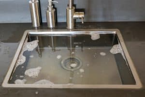 A kitchen sink filled with soapy water, bubbles gently floating on the surface. The stainless steel faucet and soap dispensers add a sleek touch, expertly installed by a plumber from Plymouth County, MA.