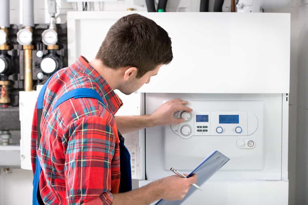 A technician in a red plaid shirt and blue overalls adjusts the settings on a white boiler in a Bristol, MA utility room, holding a clipboard and pen while checking the controls and display.