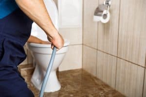 A person in a blue uniform from a plumbing contractor in Plymouth County, MA, uses a plumber's snake to unclog a toilet. The bathroom features beige tiles and a marble floor, with a roll of toilet paper visible on the wall.