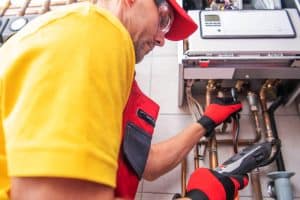 A person wearing a yellow shirt, red cap, and gloves, presumably a plumber from Plymouth County MA, is inspecting a boiler system with a diagnostic tool in the utility room. Copper pipes are visible around the unit.
