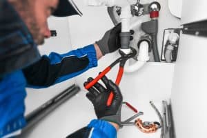 A plumber Plymouth County MA, wearing gloves and a cap, uses red-handled pliers to work on pipes under a sink. Various tools are scattered nearby in this focused scene of plumbing repairs in a small, enclosed space.