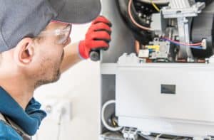 A technician wearing safety glasses and a cap is inspecting an open HVAC unit. He holds a flashlight in his gloved hand, closely examining the machine's components. Wires and parts are visible inside the unit.