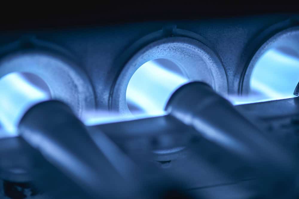 Close-up of blue flames inside a gas furnace burner in Norfolk and Suffolk County, showing metal pipes and glowing combustion chambers illuminated by the flames.