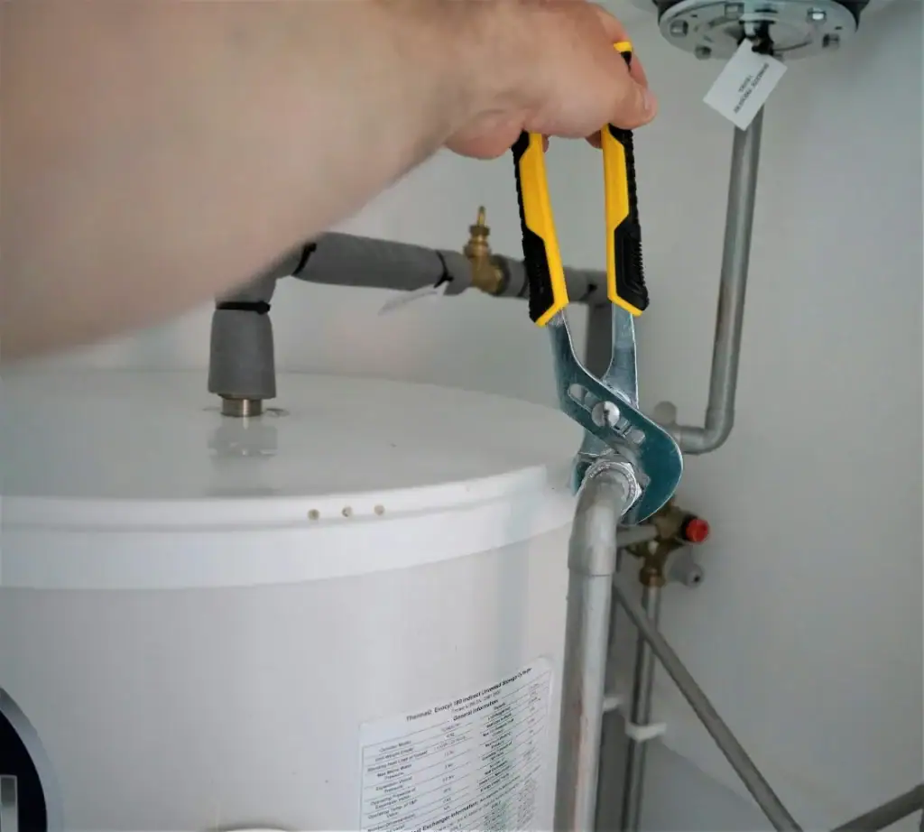 A plumber Plymouth uses adjustable pliers to tighten a pipe connected to the top of a water heater, surrounded by various plumbing pipes and fittings.