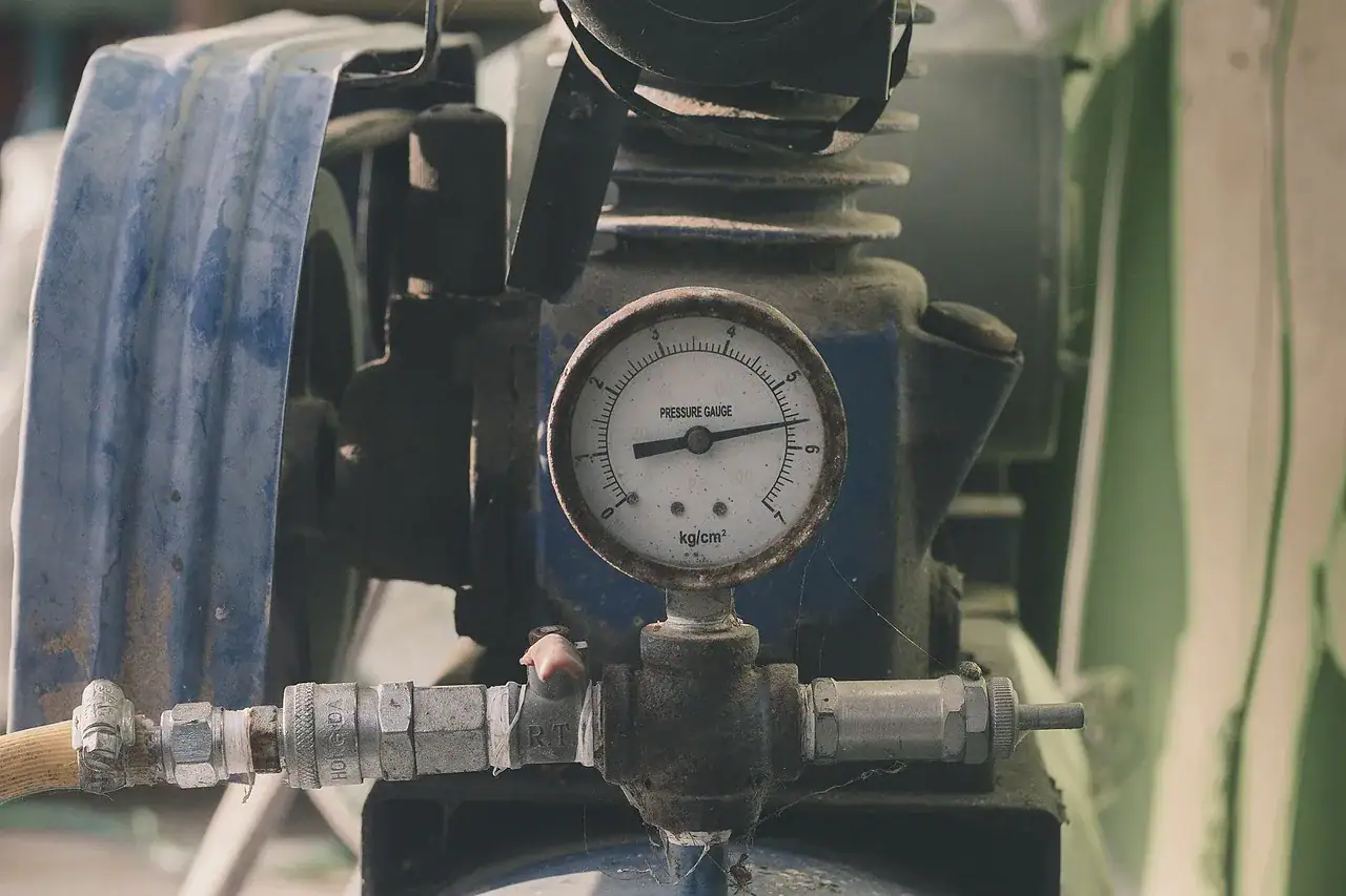 A close-up of a pressure gauge attached to industrial machinery in Bristol and Suffolk County, MA, showing measurement units. The gauge connects to pipes and fittings, with dust and signs of use visible on the equipment.