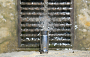 Close-up of a metal pipe leaking water forcefully in front of a drainage grate