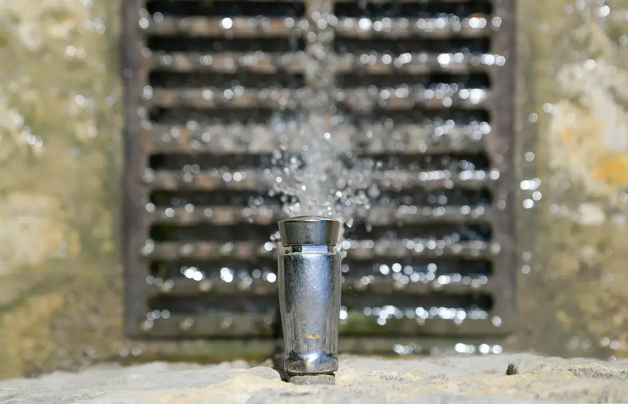 A drinking water fountain with water spraying up from the spout, set in front of a metal grate and stone background in Norfolk, MA. The fountain is in sharp focus, while the background is blurred.