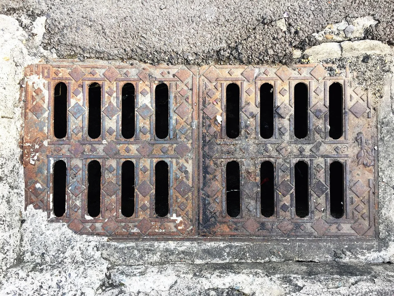 A close-up view of a rusty metal storm drain cover with rectangular slots and a geometric pattern, set in weathered concrete and surrounded by asphalt—a familiar sight for any plumber in Plymouth, Norfolk, or Middlesex.