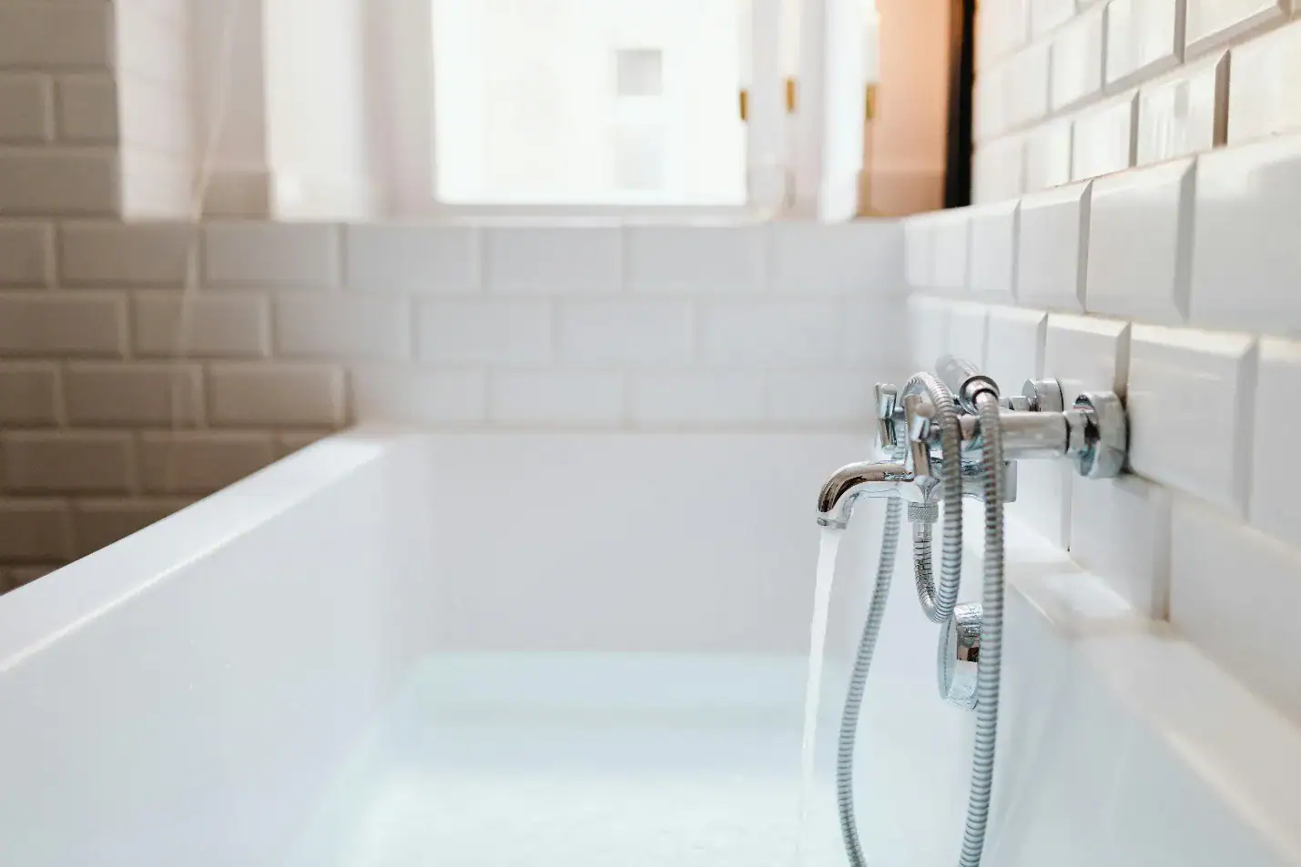 A modern white bathtub in Norfolk is being filled with water from a chrome faucet. The walls are covered with white subway tiles, and natural light enters through a nearby window.