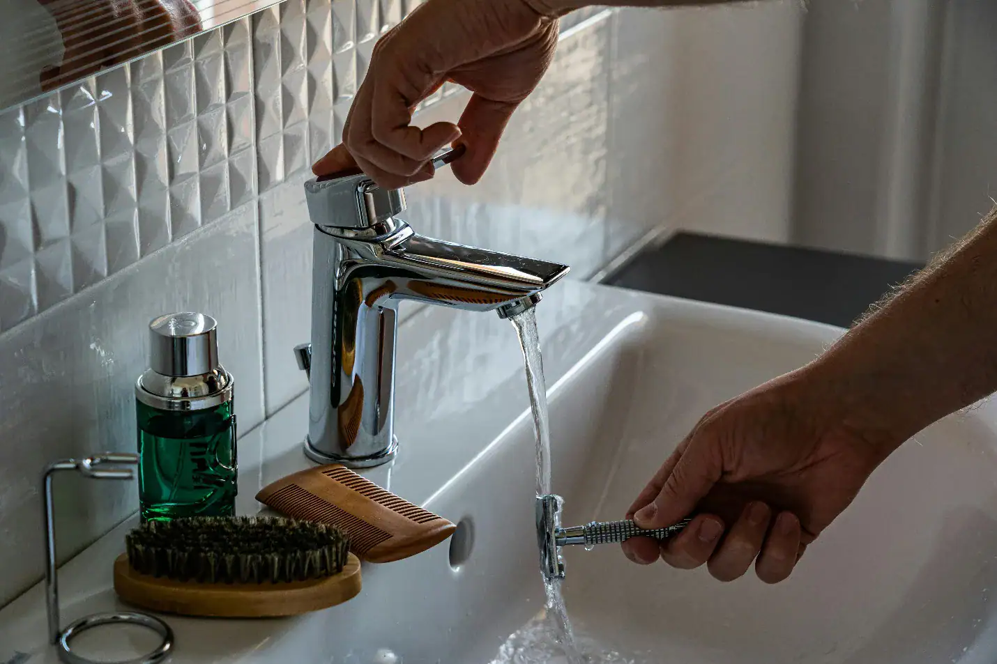 A person holds a razor under running water from a bathroom sink faucet in Norfolk. Nearby, a green bottle, wooden comb, and brush rest on the white sink countertop.