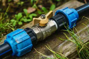 A close-up of a brass water valve with a yellow handle, connected to black plastic pipes using blue connectors, lies on a wooden surface surrounded by grass and plants—typical work for a skilled plumber Plymouth.