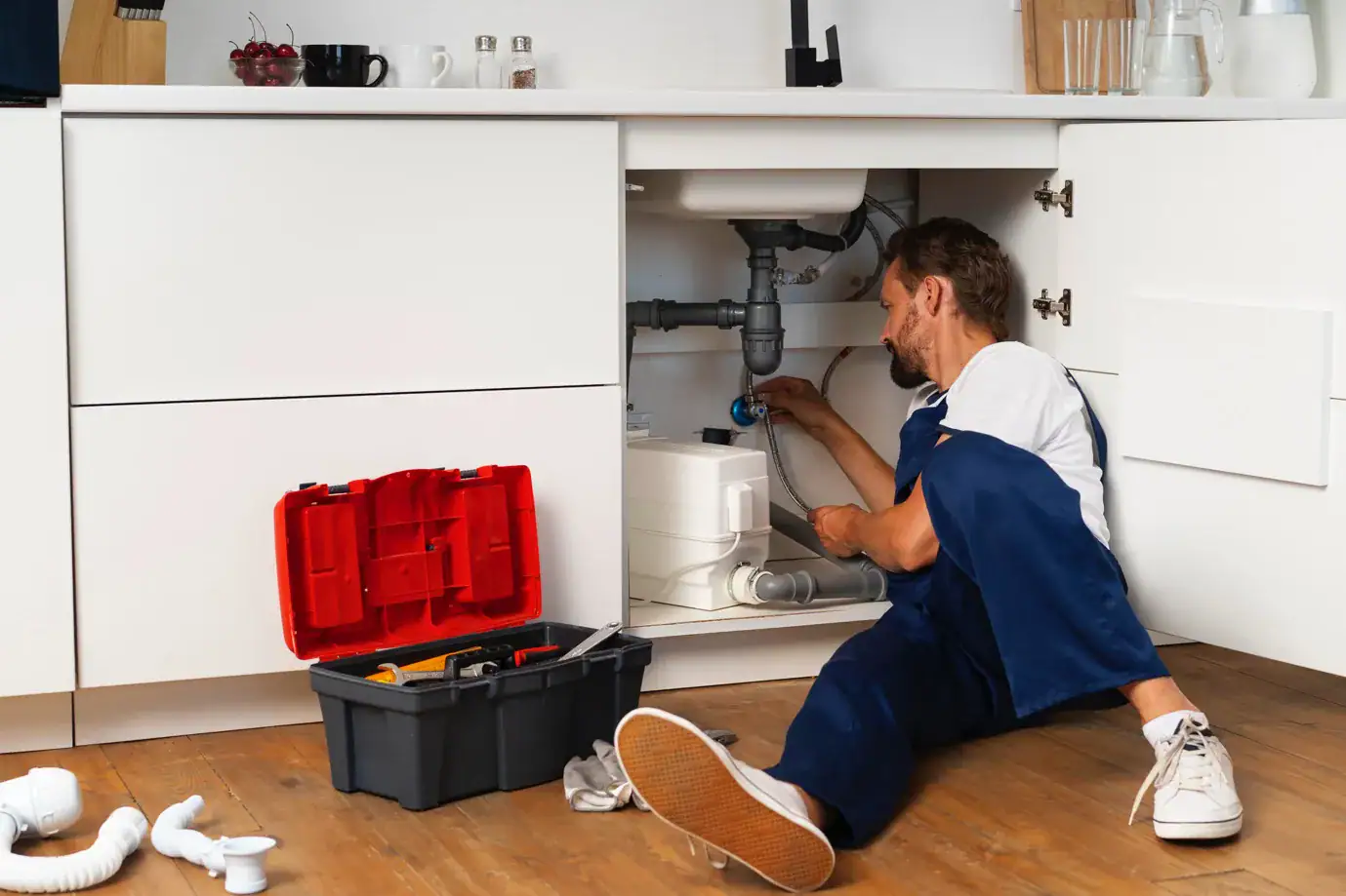 A plumber in overalls sits on the floor fixing pipes under a kitchen sink in Middlesex, MA, surrounded by a toolbox and tools, with cabinet doors open.