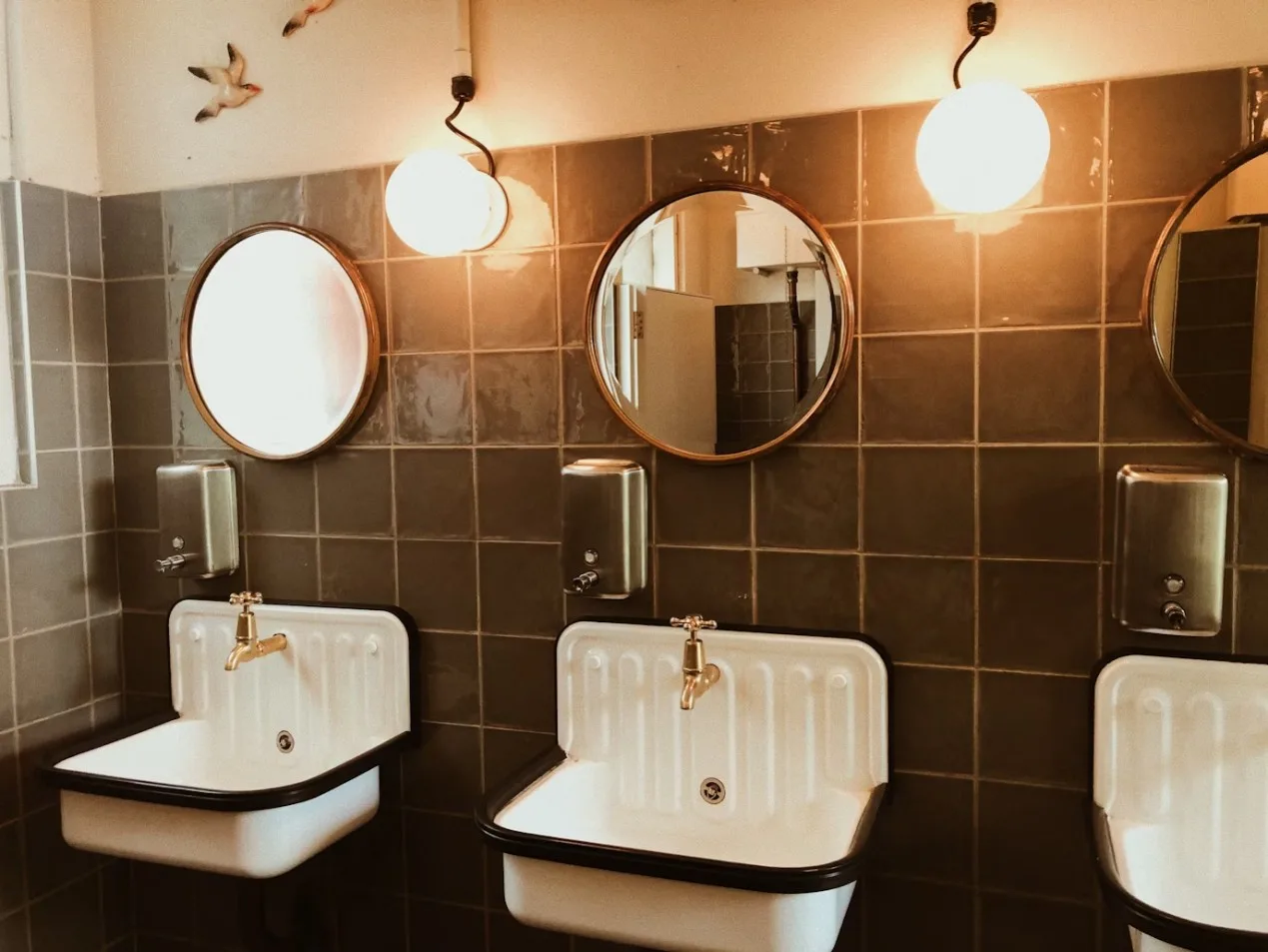 Two white sinks with brass faucets are below round mirrors and globe lights in a tiled bathroom. Soap dispensers are mounted above the sinks, and two bird decorations adorn the wall—perfect for homes in Bristol and Suffolk County, MA.