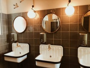 Three white sinks with gold faucets are mounted on a tiled wall in this Norfolk, MA space, each below a round mirror and globe light. Soap dispensers rest above each sink, while two decorative birds adorn the upper left wall—crafted by a skilled plumber Plymouth trusts.