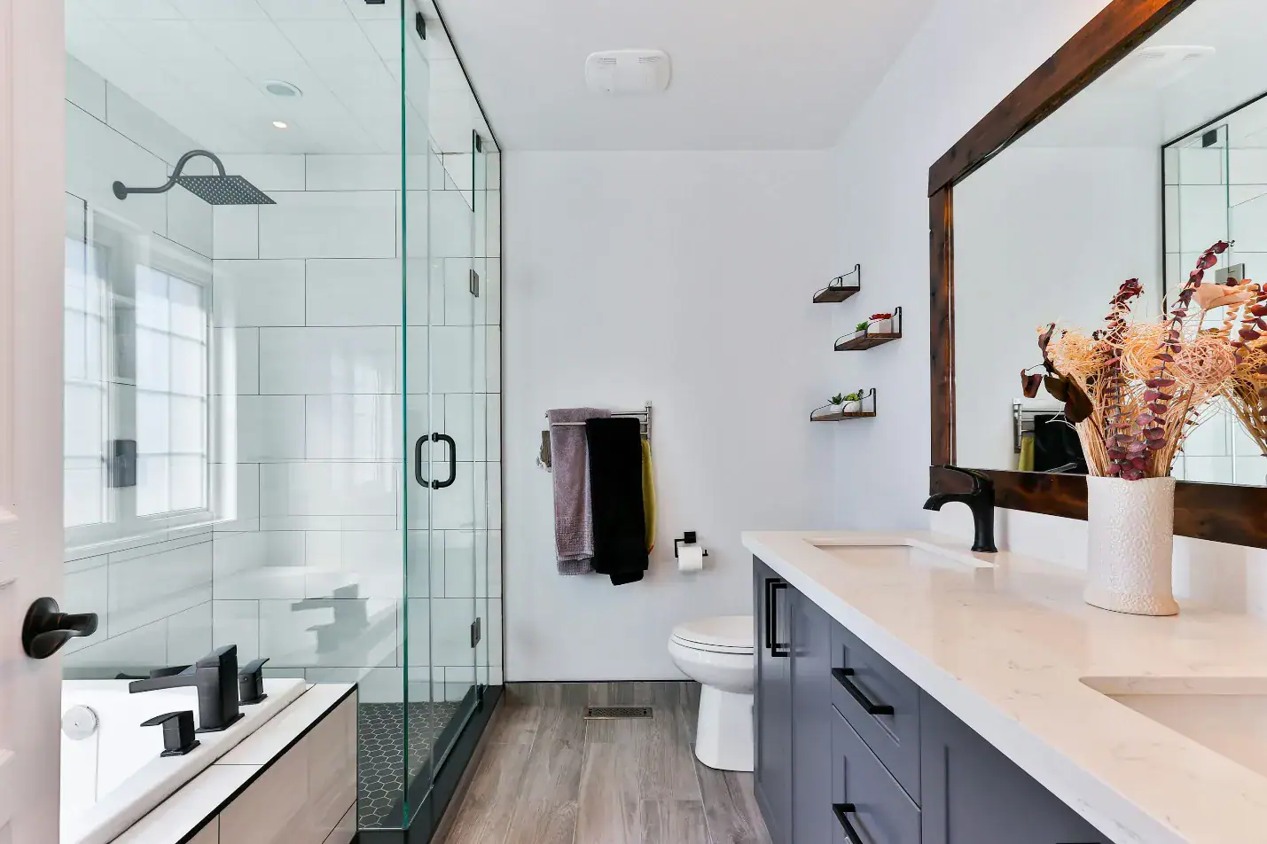 Modern bathroom in Bristol with a glass-enclosed shower, bathtub, double sink vanity with large mirror, and a vase of dried flowers. Towels hang near the toilet, and shelves hold toiletries—expertly finished by a skilled plumber Plymouth residents trust.