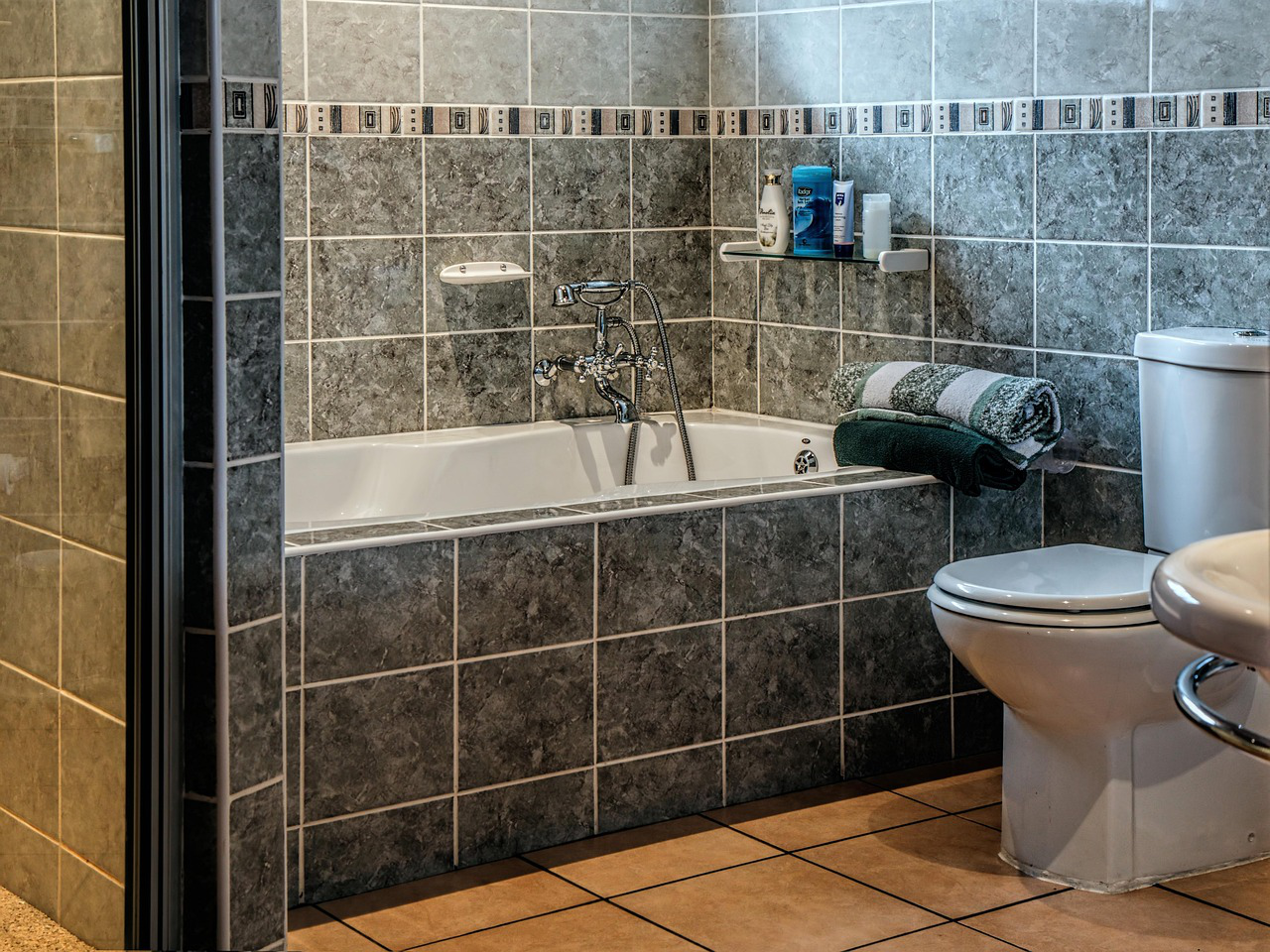 A bathrofully marbled washroom interior om with gray tiled walls and a white bathtub, equipped with a chrome faucet. Toiletries are on a shelf above the tub, and a toilet sits nearby. Towels are folded on the bath edge. The floor is tiled in brown.