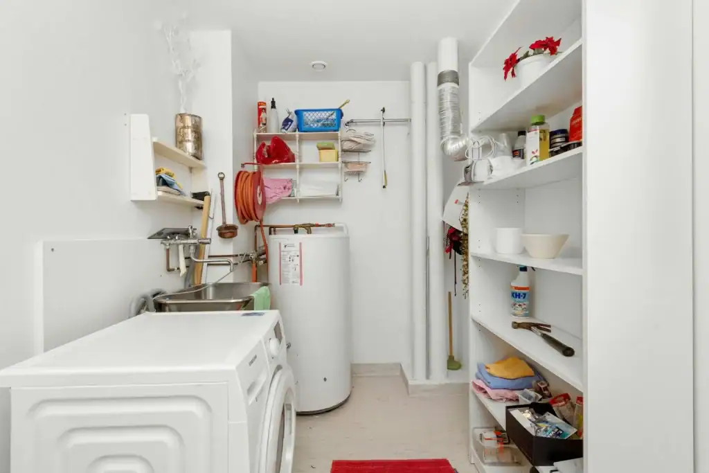 A bright laundry room in Bristol and Suffolk County with a washing machine, water heater, wall-mounted sink, shelves holding cleaning supplies and household items, and pipes running vertically along the back wall—perfect for a plumber Plymouth inspection.