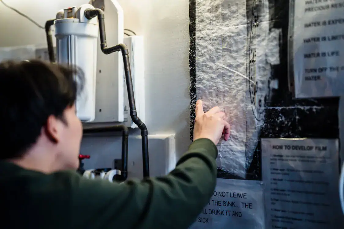 A person points at instructions posted on a wall inside a room with various pipes and equipment nearby, highlighting notes about water use and film development—perhaps referencing guidance from a plumber Plymouth serving Middlesex and Suffolk County.