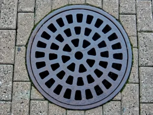 A round metal manhole cover with a pattern of rectangular openings, set into a paved surface of rectangular stone bricks in MA’s Norfolk and Suffolk County, with some green moss visible along the edge.
