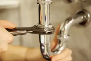 Close-up of a person’s hands using a wrench to tighten a shiny metal pipe under a sink, with a blurred background—ideal for plumbing projects in Norfolk and Suffolk County.
