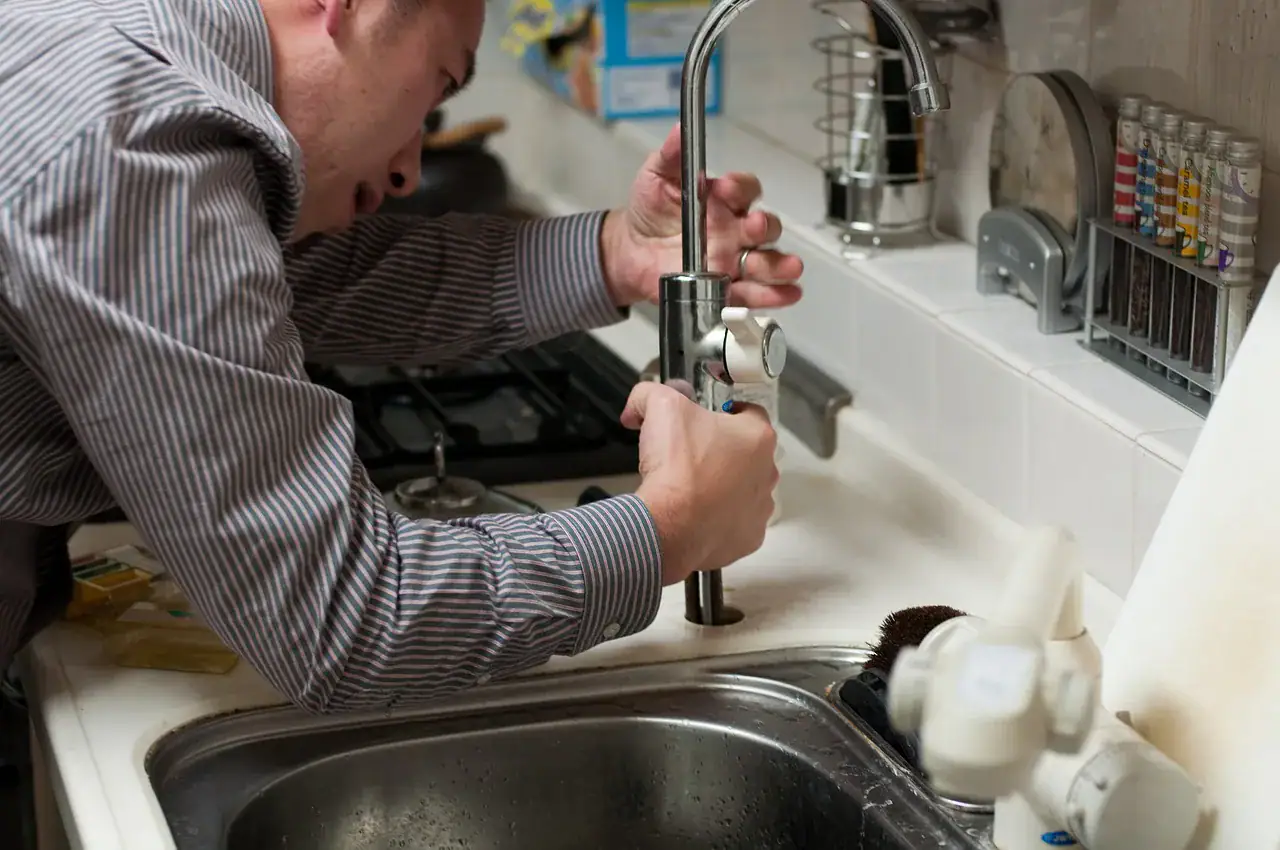 A person in a striped shirt, possibly a plumber from Plymouth or Norfolk, is repairing or installing a kitchen faucet, leaning over the sink with various tools and household items visible on the countertop.