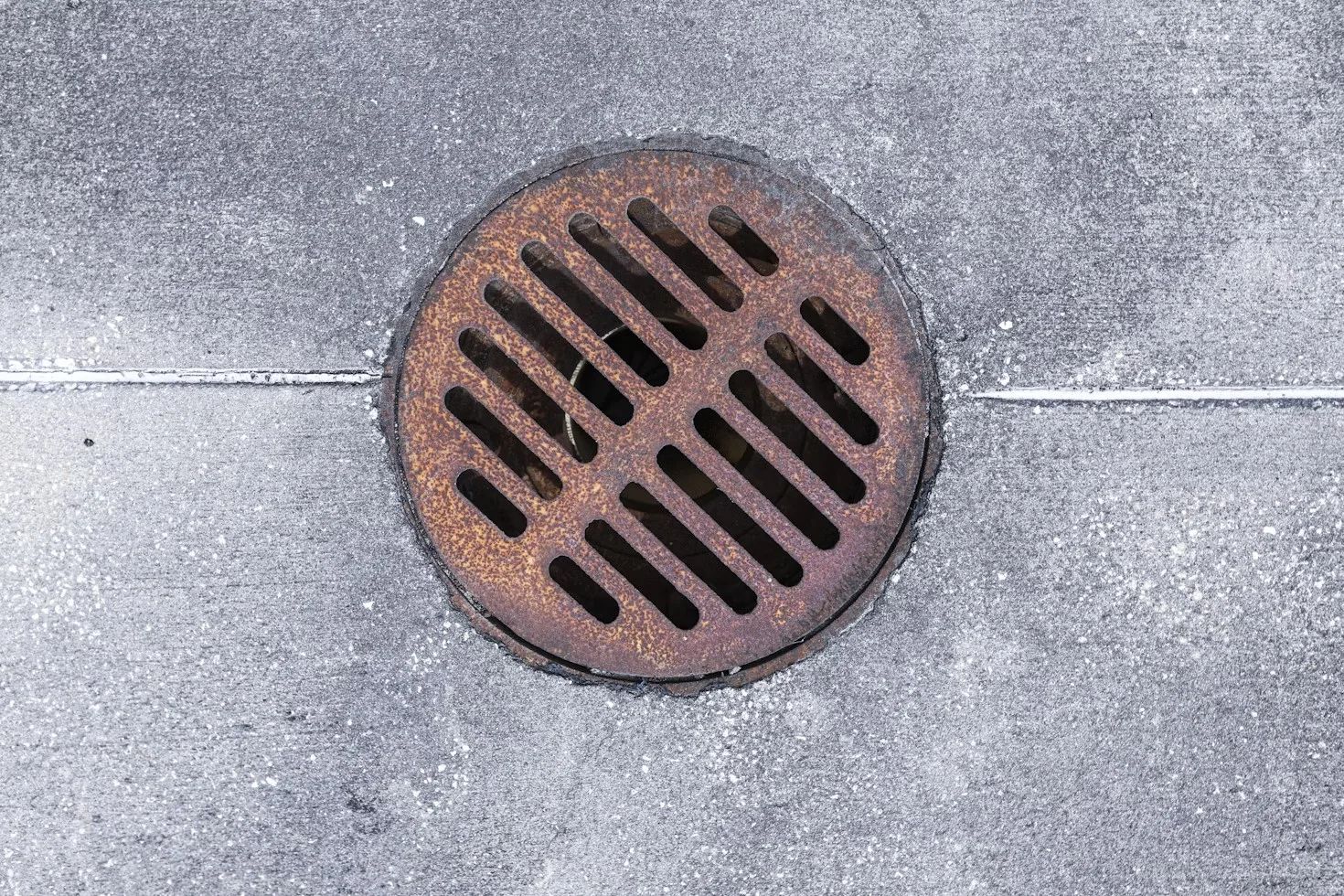 A round, rusty metal drain cover with diagonal slats is set into a gray concrete surface with visible seams and slight texture—an everyday sight for any plumber in Plymouth and Suffolk County, MA.