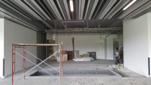 A large, unfinished indoor space in MA with striped ceiling panels, exposed wiring, construction materials scattered on the floor, scaffolding in the foreground, and two workers—possibly a plumber from Plymouth or Bristol—near the back wall.