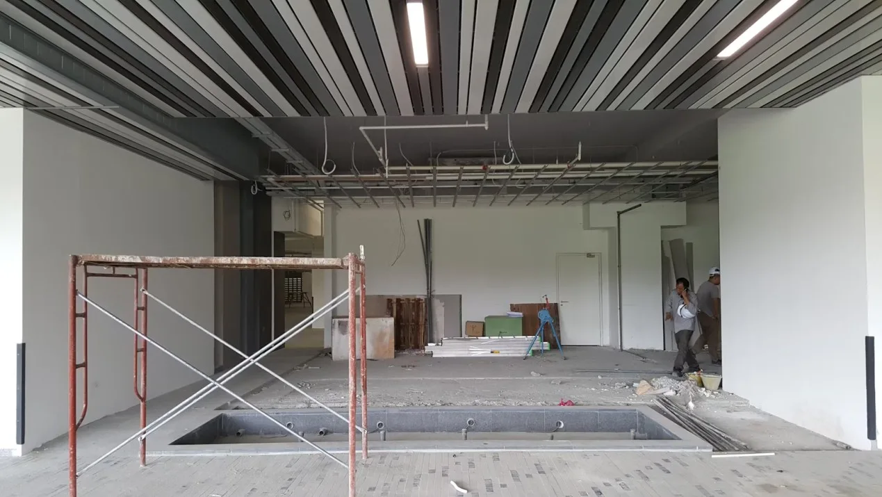 A large, unfinished indoor space in MA with striped ceiling panels, exposed wiring, construction materials scattered on the floor, scaffolding in the foreground, and two workers—possibly a plumber from Plymouth or Bristol—near the back wall.