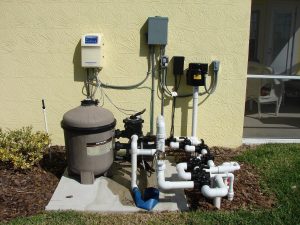 Outdoor pool pump and filtration system with multiple white PVC pipes, control boxes, and a round filter unit installed on a concrete pad next to a yellow stucco wall. For expert setup in MA, contact a trusted plumber Plymouth residents recommend.